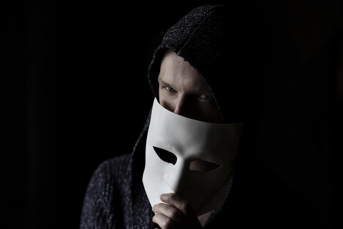 Fotografia di una persona incappucciata che si copre parzialmente il volto con una maschera bianca, a rappresentare l'anonimato di chi compie attacchi informatici